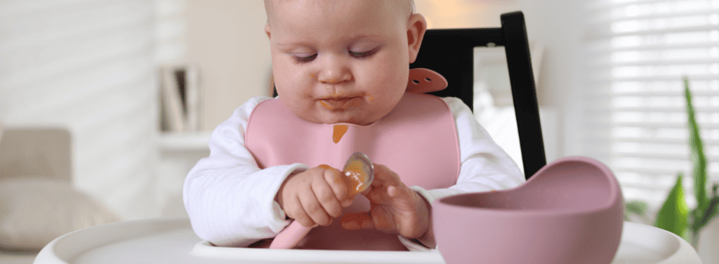 Baby sat in highchair eating with a spoon